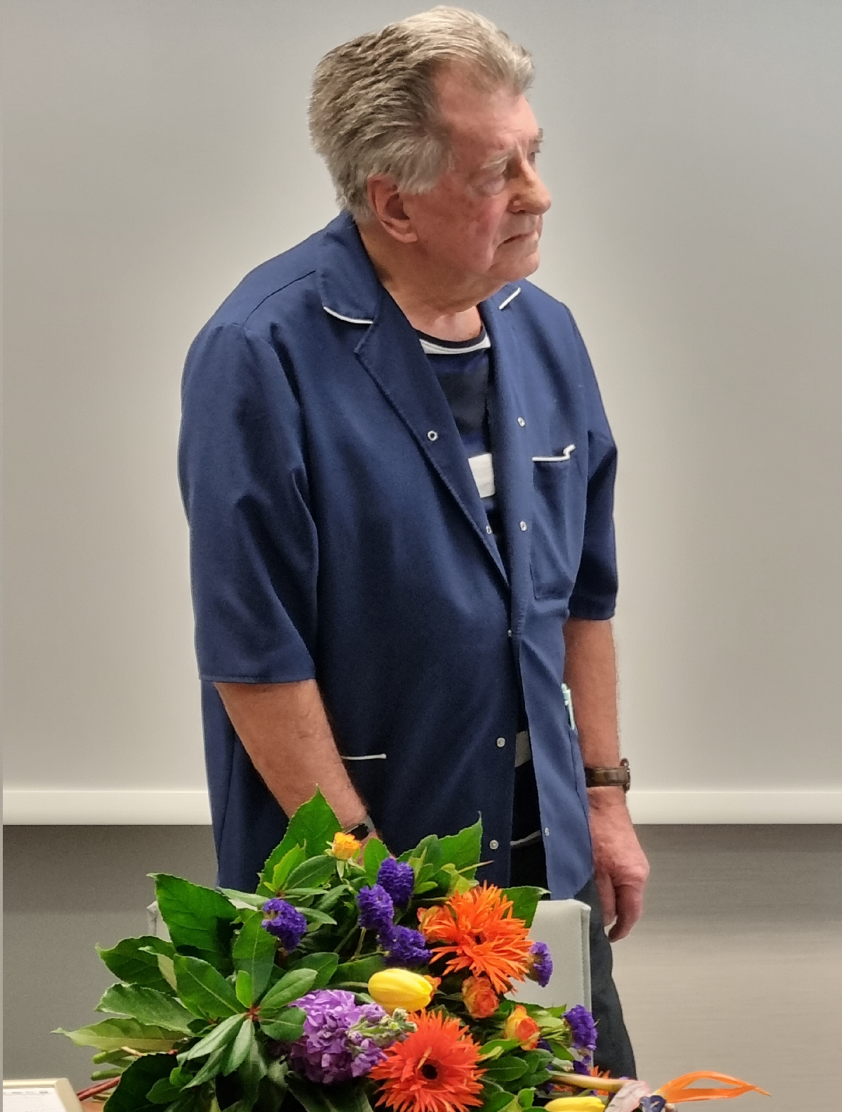 Starszy mężczyzna w niebieskiej koszuli i pasiastym podkoszulku, stojący obok bukietu kwiat&oacute;w, patrzy w prawo.